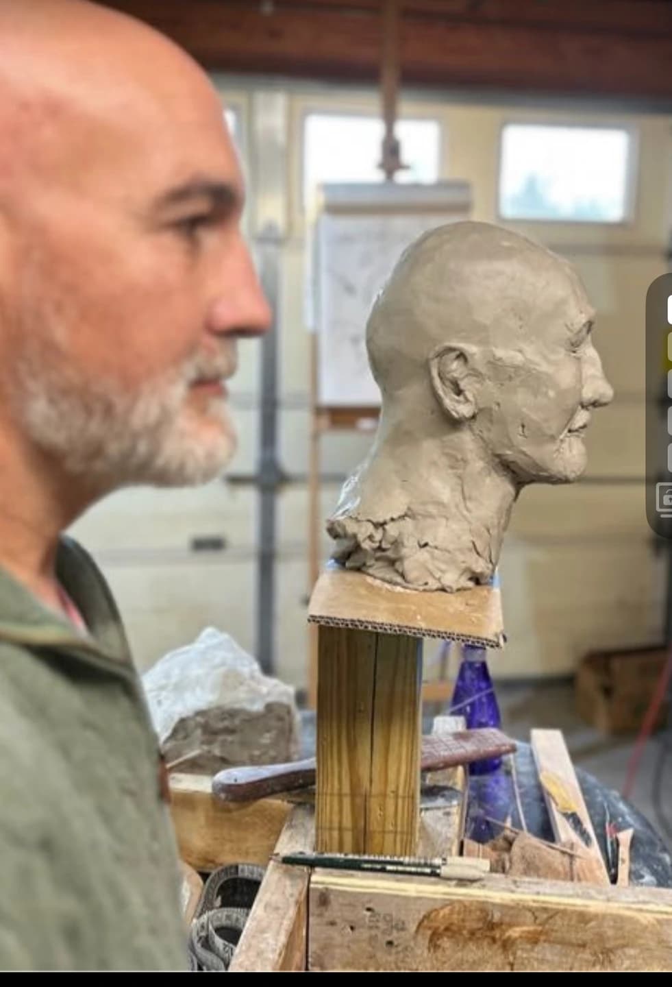 A person in profile beside a clay portrait bust and sculpting tools.